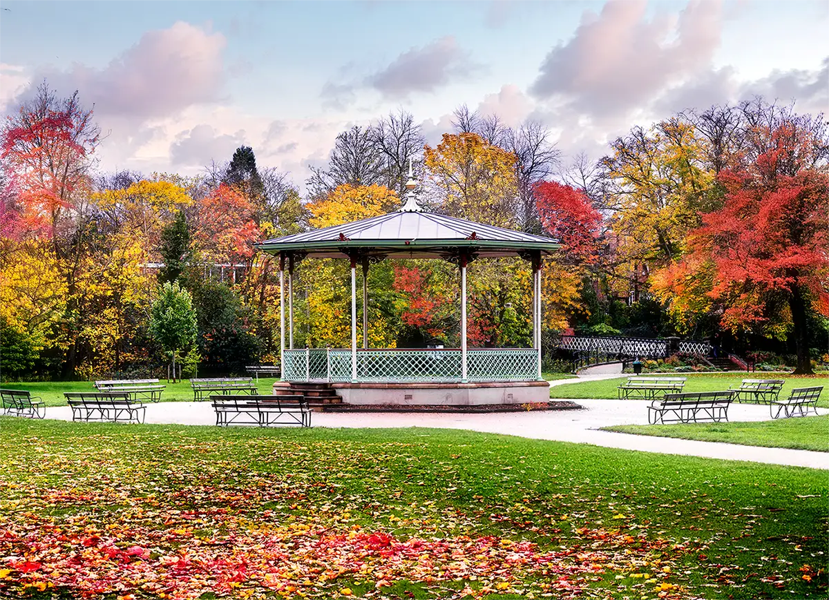 A daytime photograph of the historic Victorian bandstand in the Pump Room Gardens, Royal Leamington Spa, surrounded by vibrant autumn foliage, park benches, and fallen leaves scattered on the grass.