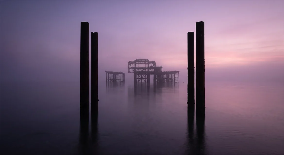 Fine art photography of the iconic iron architecture of West Pier in Brighton framed by dark structural pillars, receding softly into an amethyst-toned morning mist.
