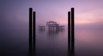 Fine art photography of the iconic iron architecture of West Pier in Brighton framed by dark structural pillars, receding softly into an amethyst-toned morning mist.