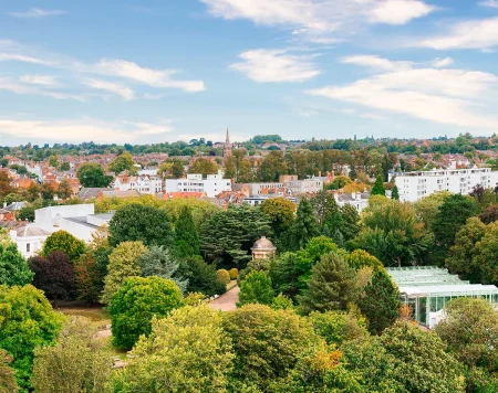 An elevated view overlooking Jephson Gardens in Royal Leamington Spa, showing the glasshouse, mature trees, and the spire of All Saints' Church on the horizon.