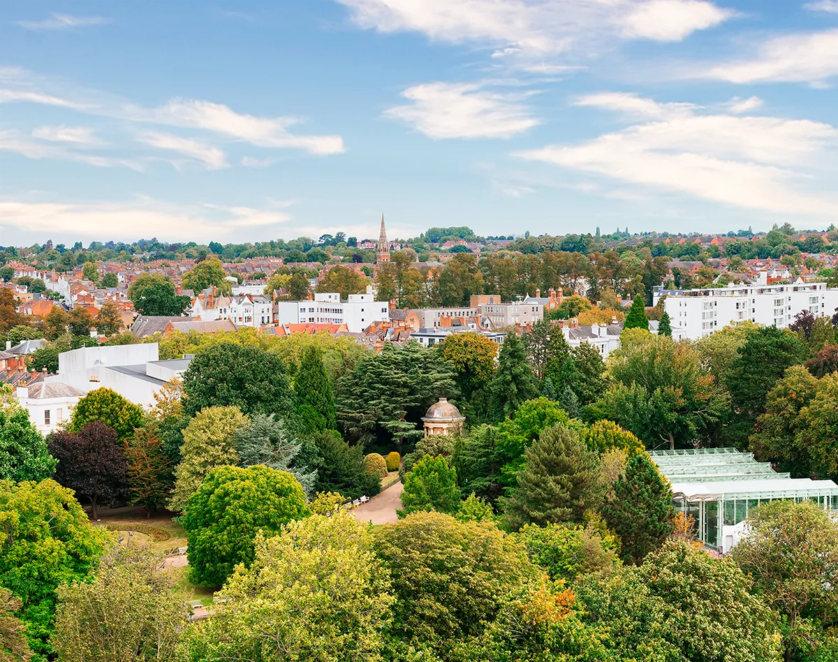 An elevated view overlooking Jephson Gardens in Royal Leamington Spa, showing the glasshouse, mature trees, and the spire of All Saints' Church on the horizon.
