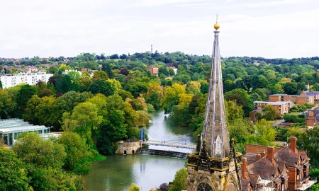A high-angle photograph looking across the River Leam in Royal Leamington Spa, featuring the stone spire of All Saints' Church, the weir, and the blue pedestrian suspension bridge.