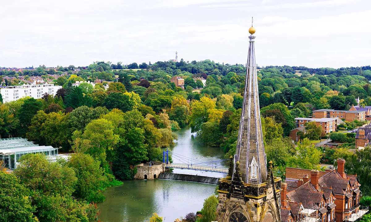 A high-angle photograph looking across the River Leam in Royal Leamington Spa, featuring the stone spire of All Saints' Church, the weir, and the blue pedestrian suspension bridge.