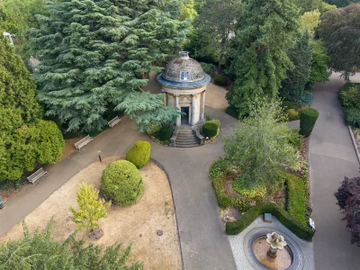 High-angle aerial view looking down at the historic stone Jephson Memorial pavilion surrounded by mature cedar trees, paved pathways, manicured topiary, park benches, and a small circular fountain in Jephson Gardens, Royal Leamington Spa.